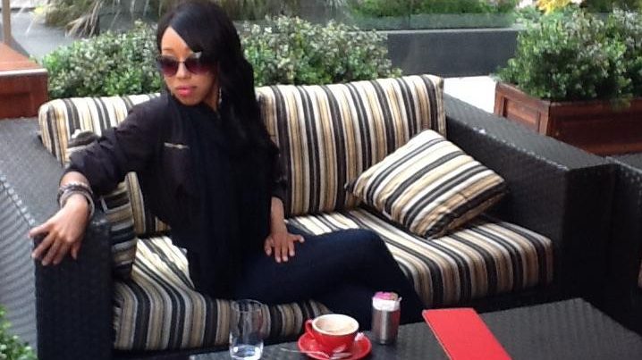 Stylish woman in sunglasses seated on a striped outdoor sofa at a city café, with coffee and a red menu on the table in front of her.